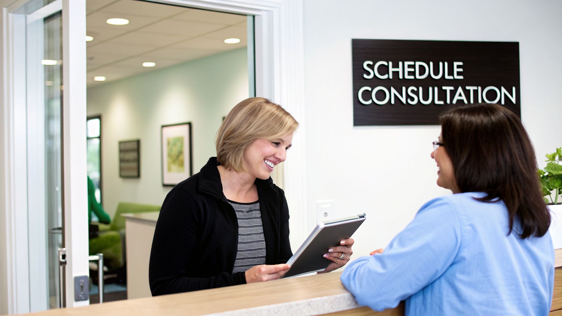 Two women smiling at a reception desk, one holding a tablet, with a 'SCHEDULE CONSULTATION' sign.