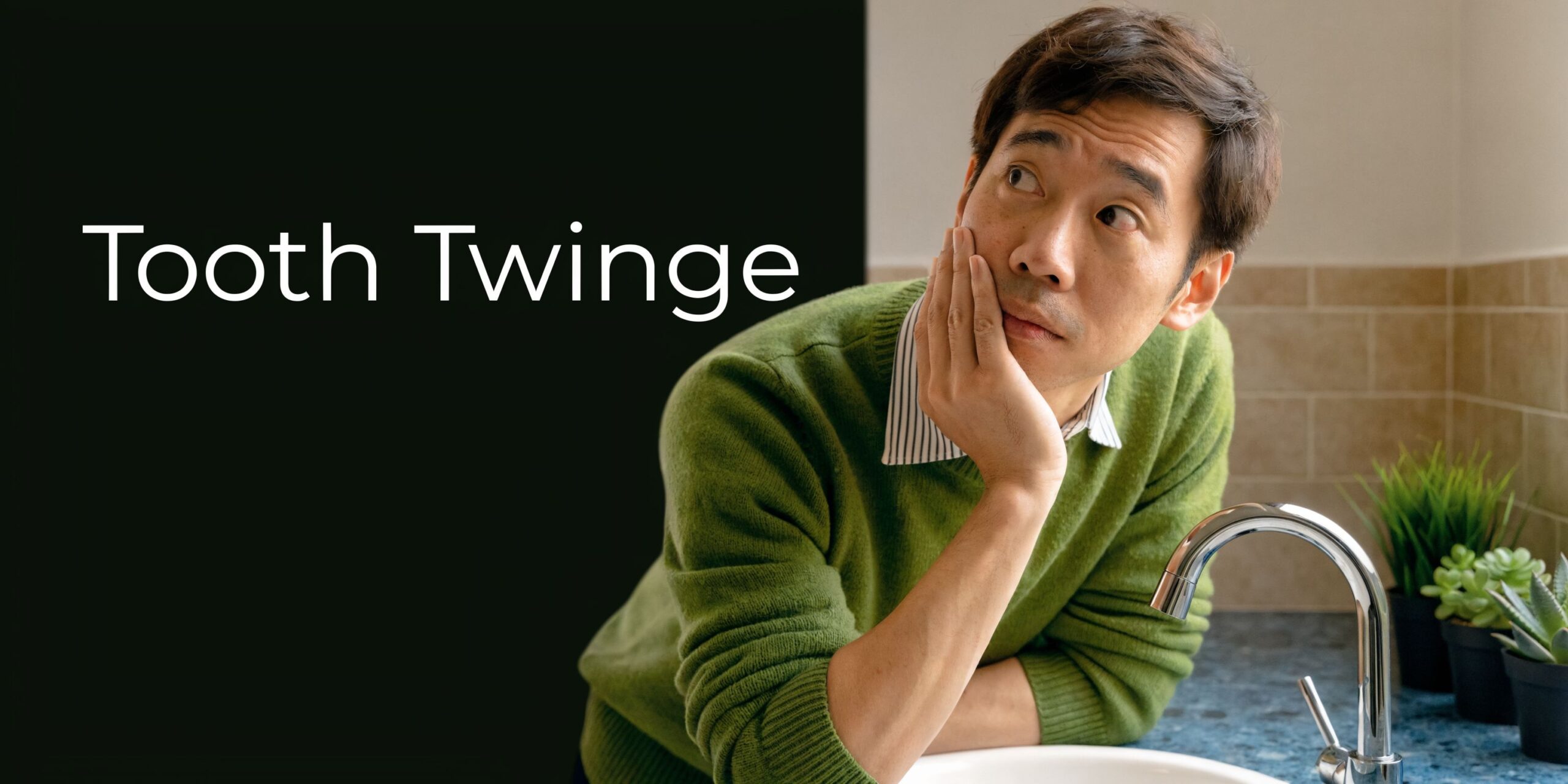 An Asian man holding his cheek in pain while standing near a bathroom sink, representing tooth sensitivity.